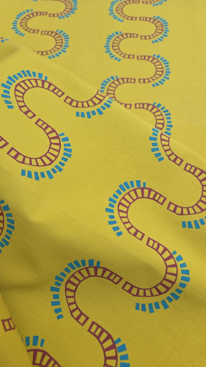 A yellow fabric featuring a repeating pattern of train tracks in a wavy design, with blue and brown rectangles representing the sleepers. The tracks are arranged in smooth curves across the fabric's surface.