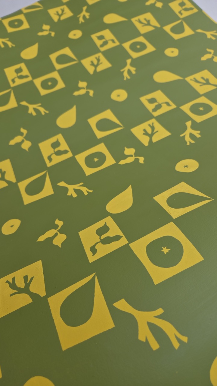 A patterned surface featuring various yellow graphic shapes, including leaves, abstract forms, and dots, arranged in a grid over a green background. The design is playful and repetitive, creating a visually interesting texture.