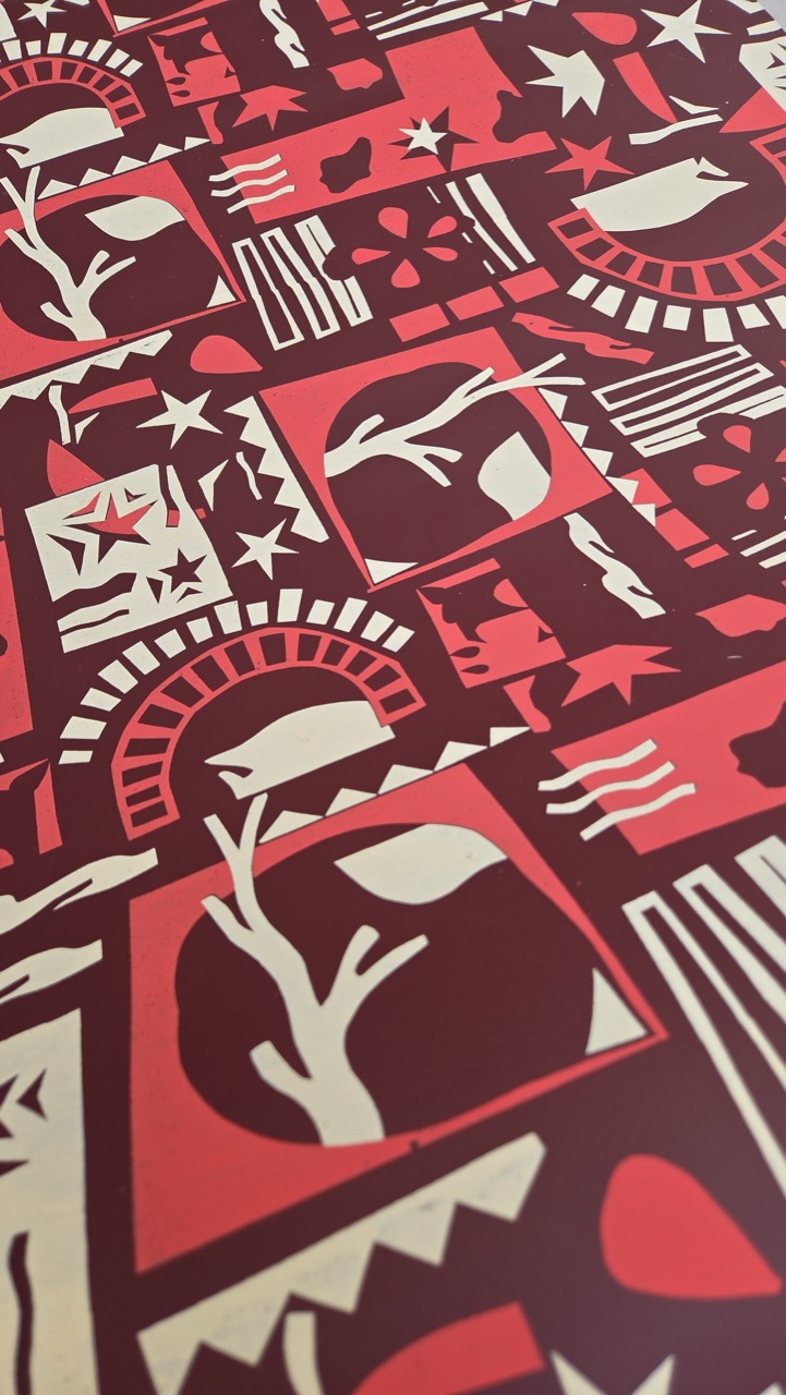 A patterned surface featuring various designs in red, cream, and dark maroon. The design includes stylised depictions of trees, birds, and stars, arranged in a grid within bordered squares.