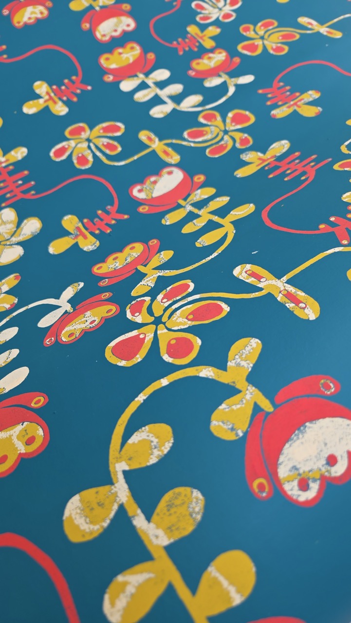 A blue background adorned with a pattern of colourful flowers and whimsical shapes. The design features red, yellow, and white floral elements with thin, curvy green stems, creating an intricate, playful visual composition.