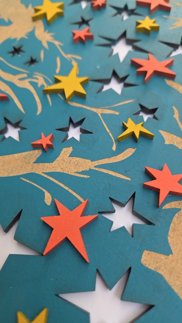 A close-up view of layered paper cutouts depicting stars in red, yellow, and white on a textured teal background. The design features branches curling among the stars, creating a visually dynamic and colourful composition.