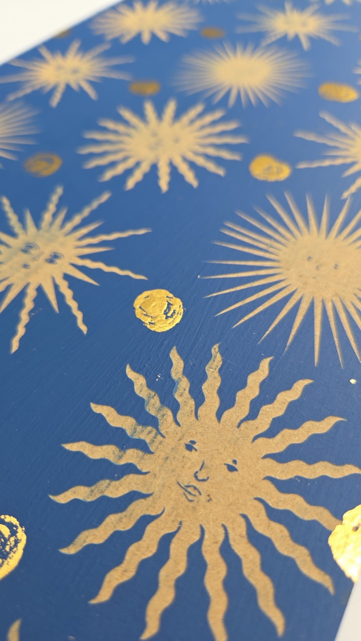 A blue surface decorated with gold sunburst patterns, featuring various sizes and styles of suns and circles arranged evenly. The design includes intricate wavy lines radiating from the centre of the suns, creating a striking visual contrast.