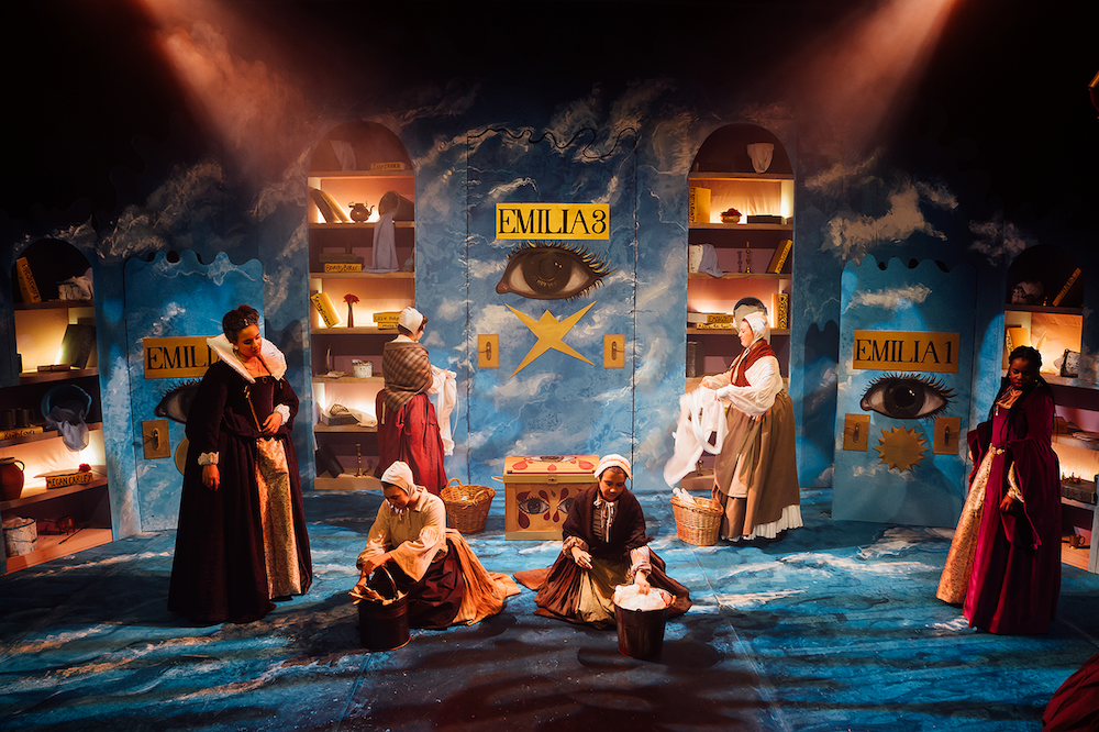 Theatre stage with blue backdrop, warm lighting, actors in period costumes, shelved set pieces with "EMILIA'S" signage, dramatic shadows.