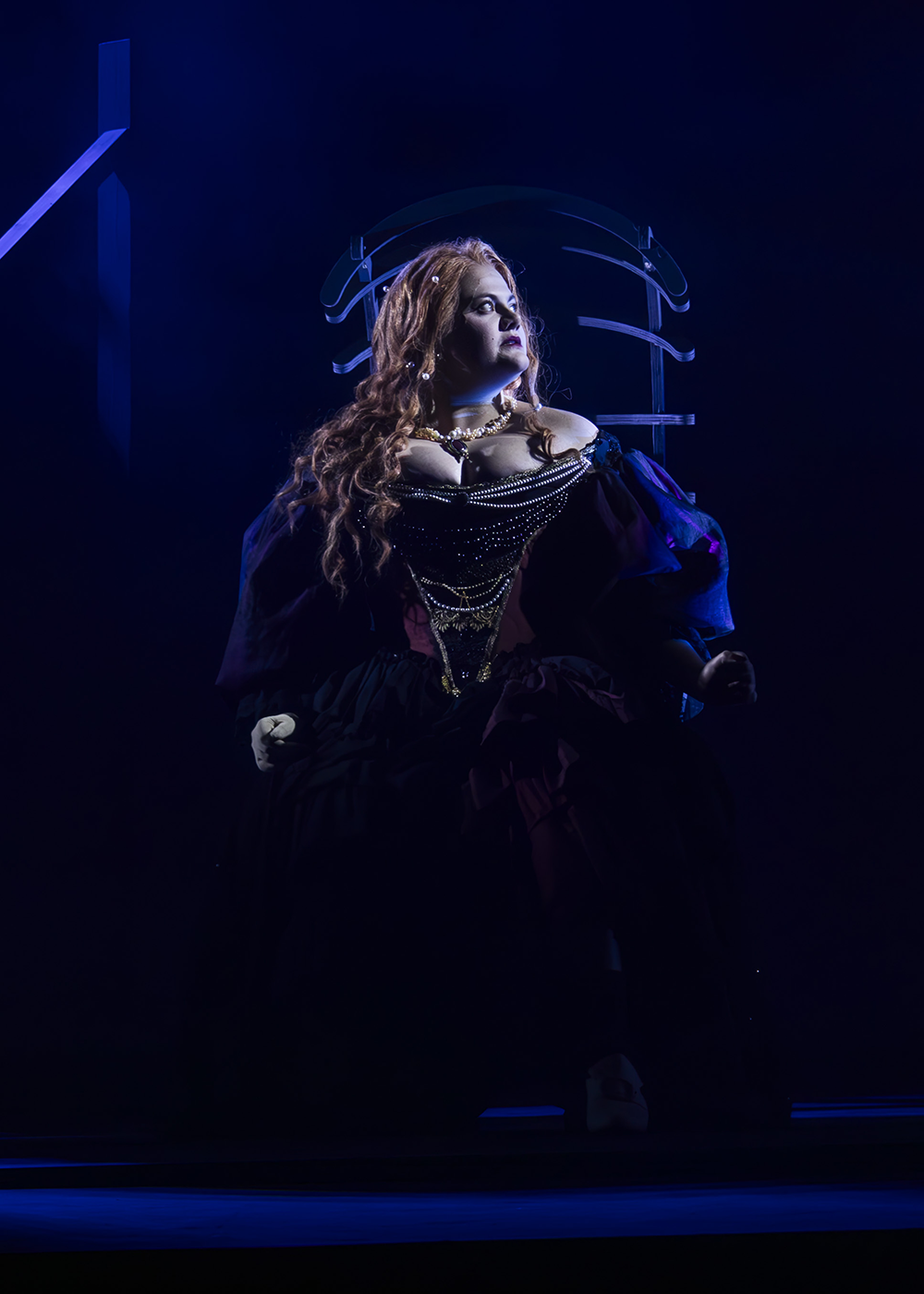Woman with long wavy hair in ornate black corset and dress, sitting in chair, blue stage lighting against dark background.