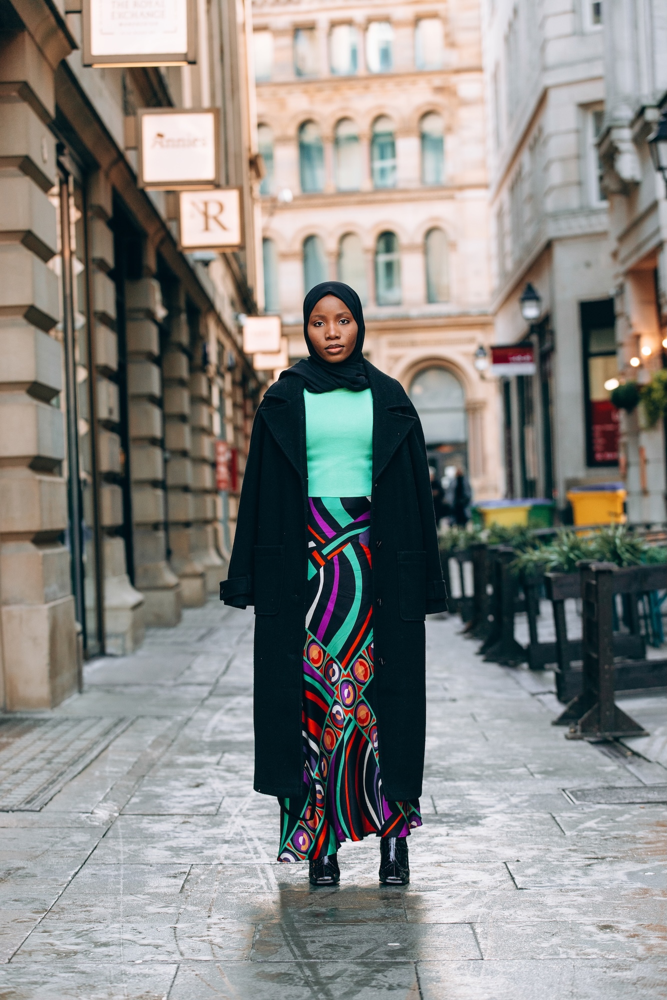 Woman in black hijab and coat with mint top and patterned maxi skirt standing on stone street between historic buildings.