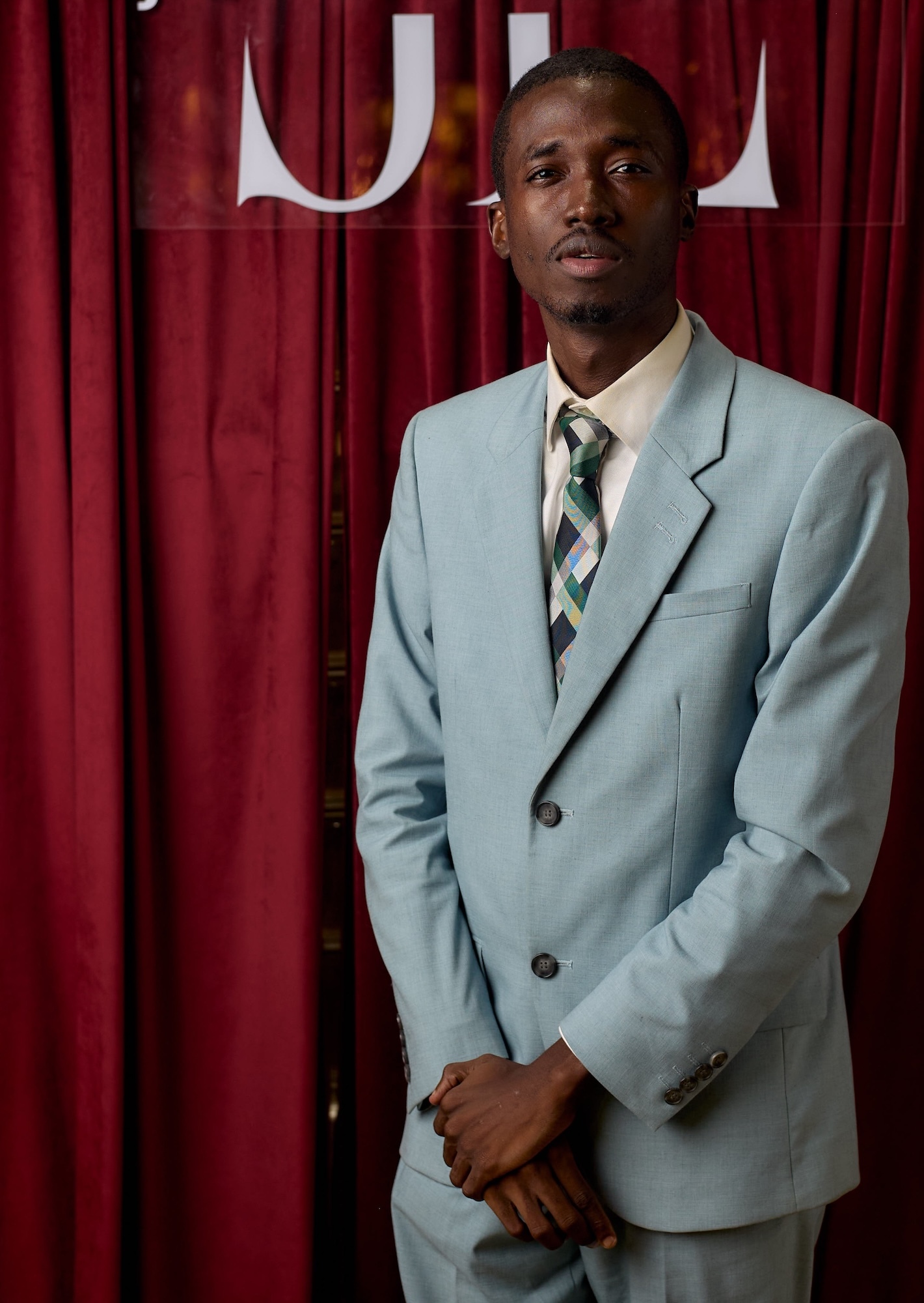 Portrait of A. Afolabi standing against red curtain backdrop with white lettering appearing above him.