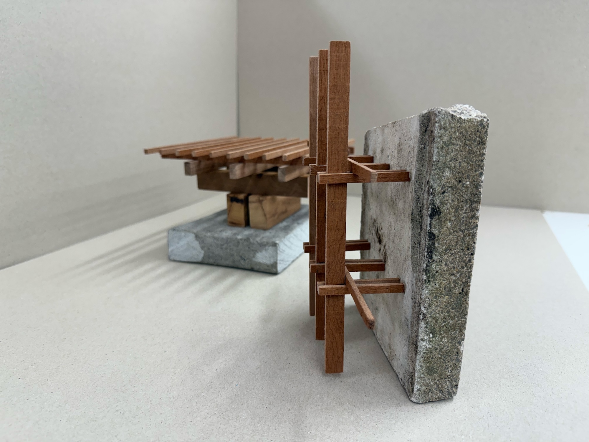Wooden architectural model with horizontal slats and vertical supports positioned against grey concrete block on white surface.