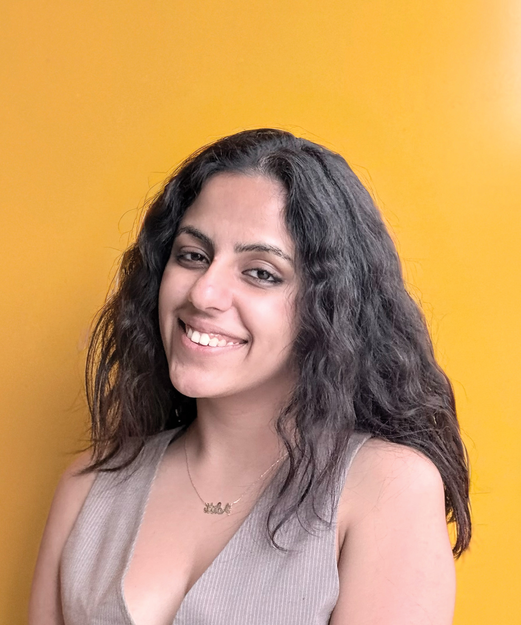 Woman with long dark wavy hair smiling, wearing beige sleeveless top, standing against bright yellow wall.