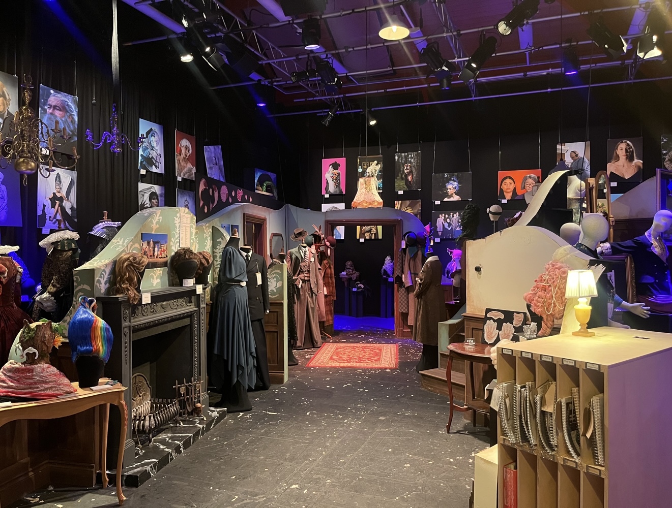 Dark exhibition space with purple lighting, displaying vintage costumes on mannequins, framed portraits on black walls, and wooden furniture pieces scattered throughout the room.