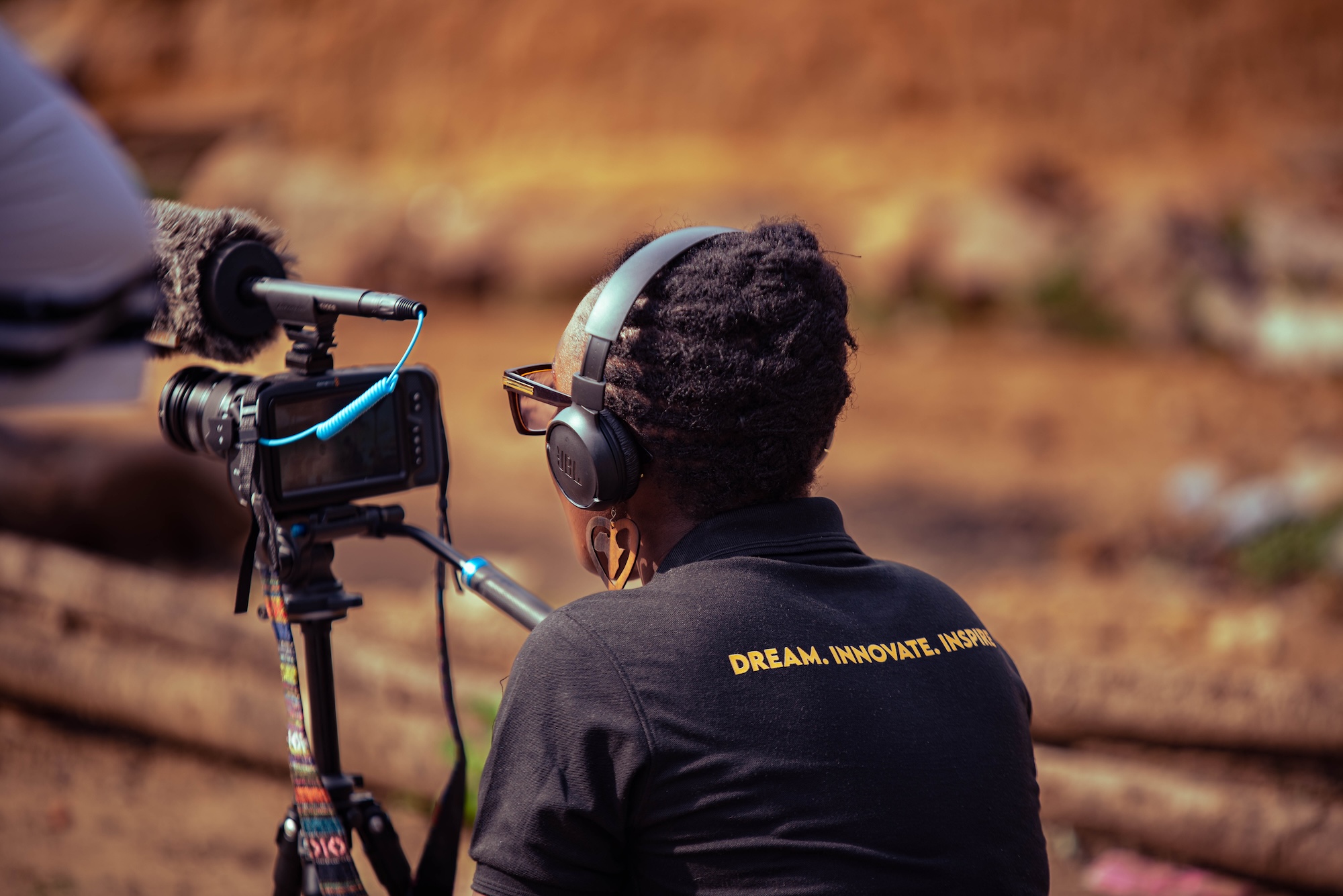 A person wearing headphones records video with a camera on a tripod, with logs and a blurred outdoor background behind them.