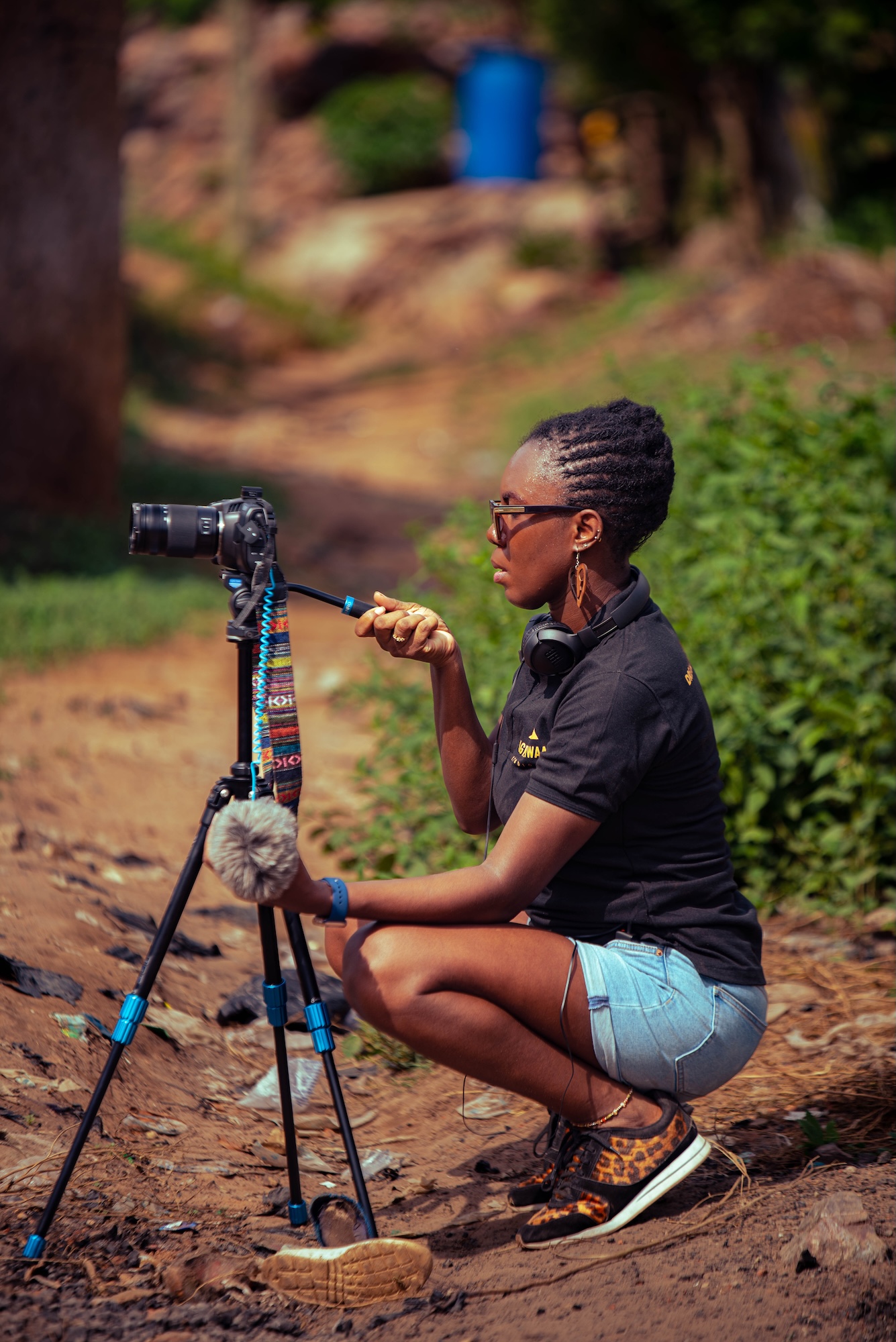 A person crouches beside a camera on a tripod in a rural path, holding a cable while wearing a black top, denim shorts and headphones.