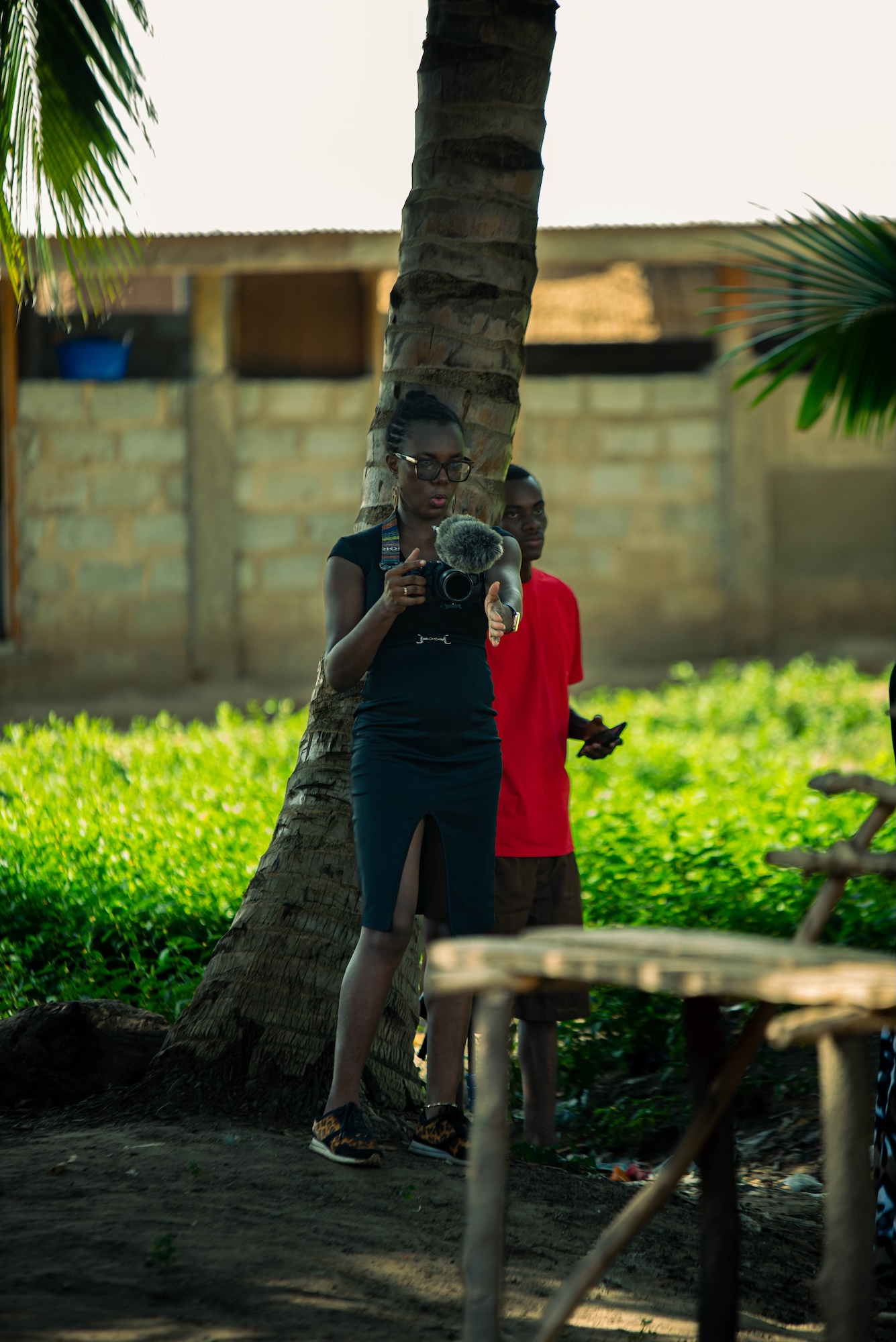 A person holding a camera stands beside a tree, with a child in a red shirt behind her and a grassy yard with a building in the background.