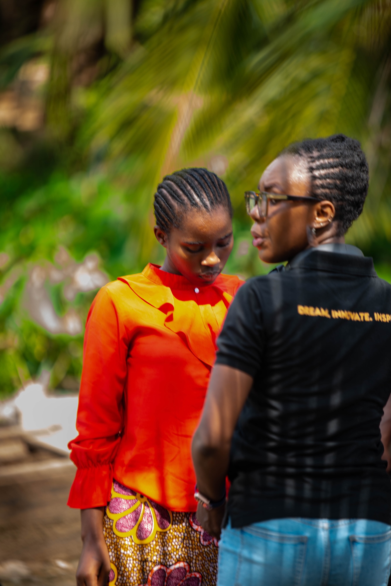 Two people stand outdoors, one in an orange blouse and patterned skirt, with palm trees and a blurred garden in the background.