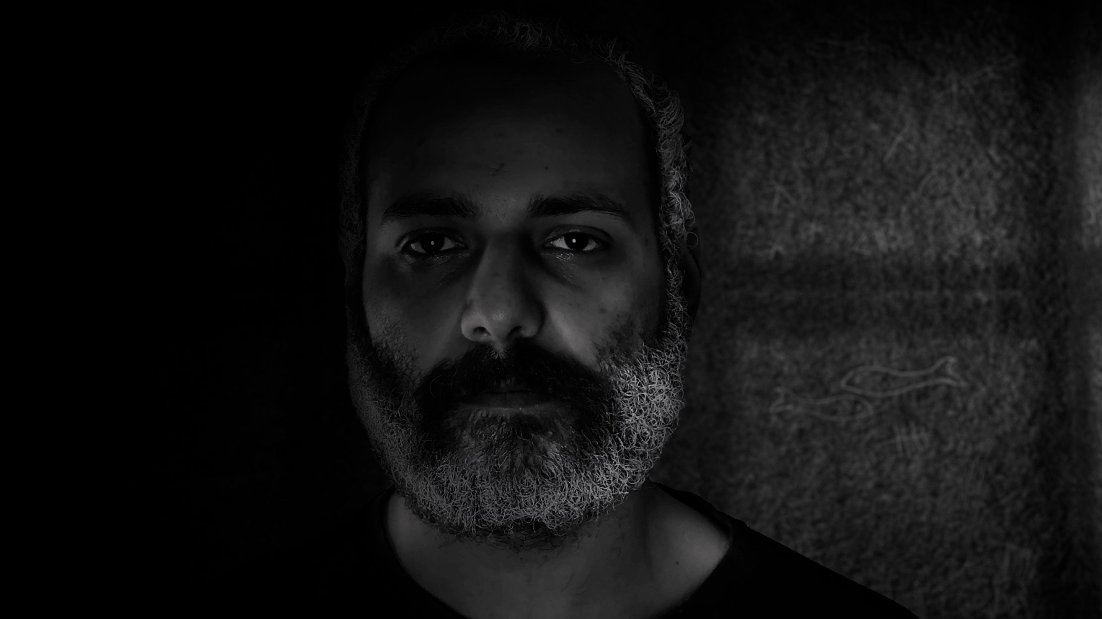 A black-and-white portrait of a bearded man in low light, with half his face in shadow against a textured background.