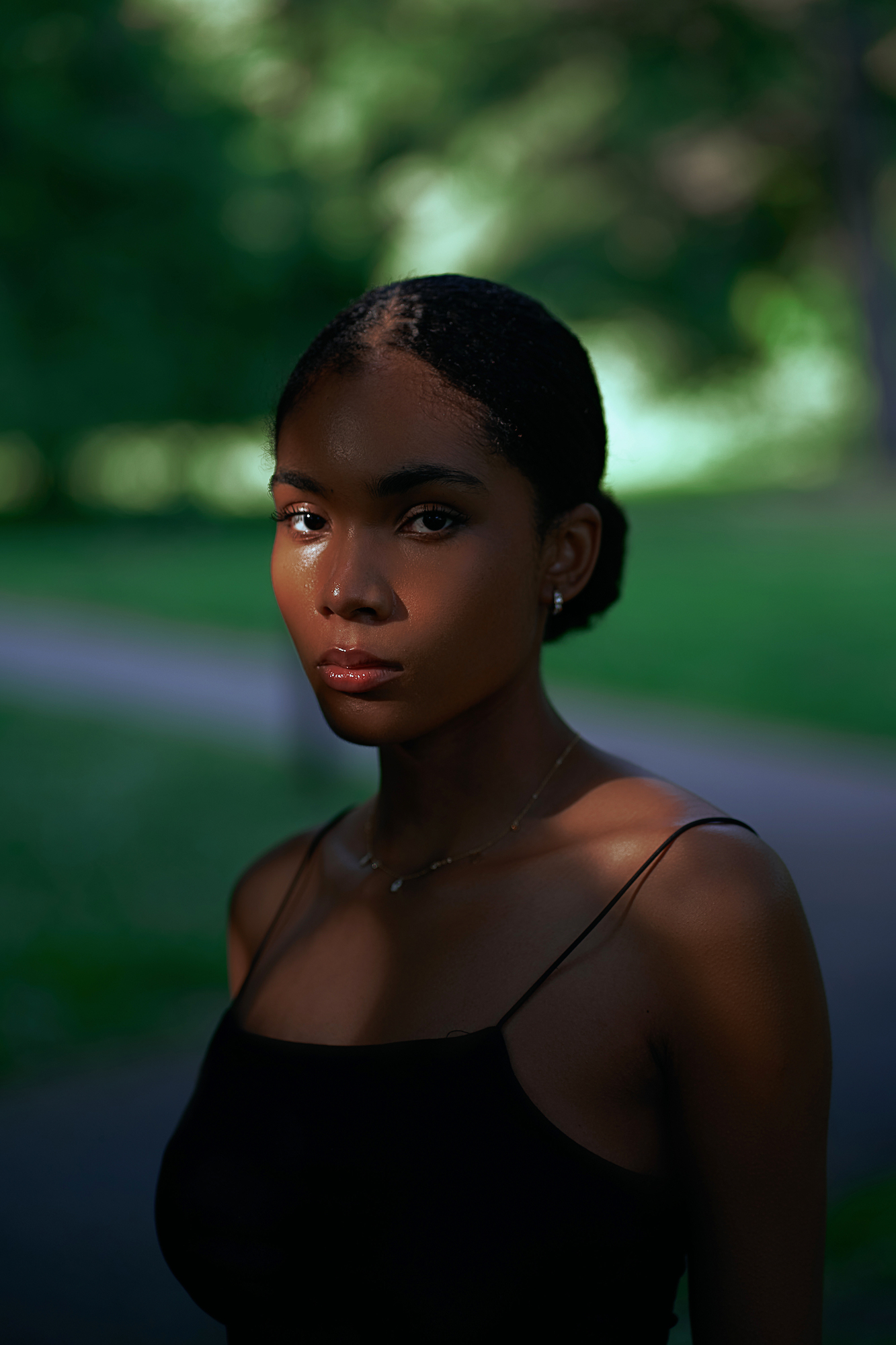Portrait of a woman looking toward the camera while standing in the shade, a footpath and grassy lawns in the background.