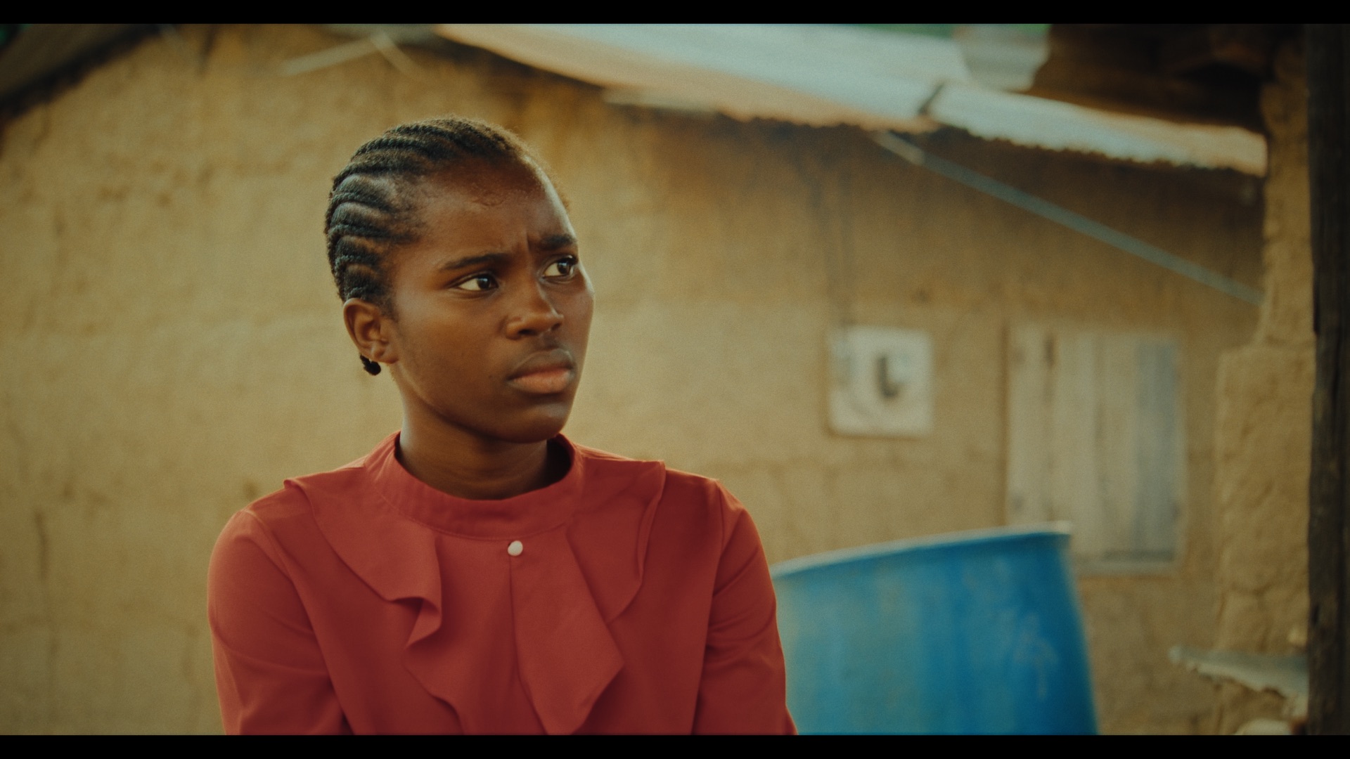 A young woman with braided hair is seated outside a building made of mud and thatch. She is wearing a red blouse with a ruffled front, and a blue water barrel is partially visible in the background.
