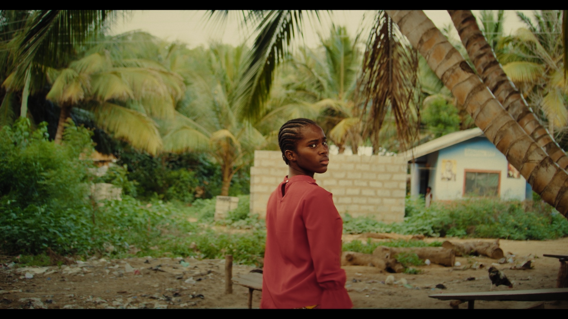 A young girl with braided hair is standing in a rural area surrounded by palm trees. In the background, there's a small blue house and some greenery, while the ground features scattered litter and a wooden bench.