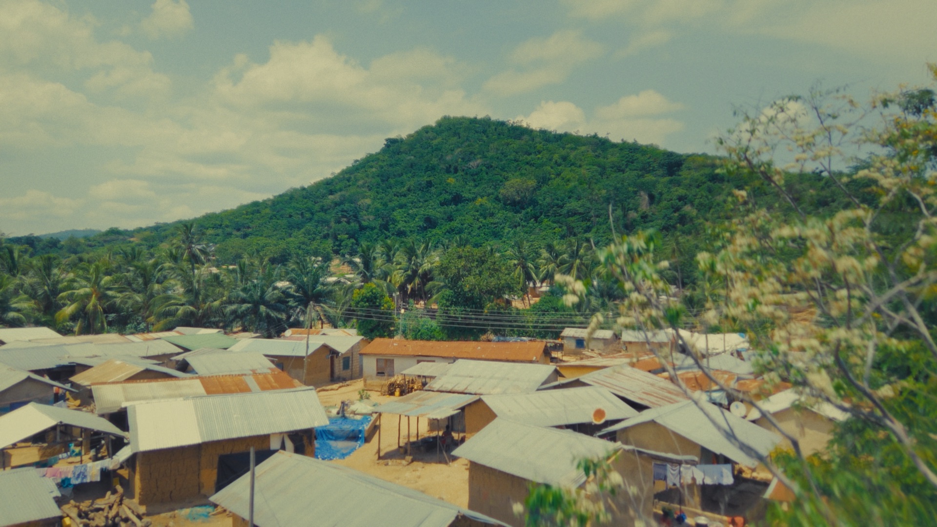 A view of a village with small houses featuring metal roofs, surrounded by lush green trees and hills. In the background, a prominent hill covered with vegetation rises against a partly cloudy sky.