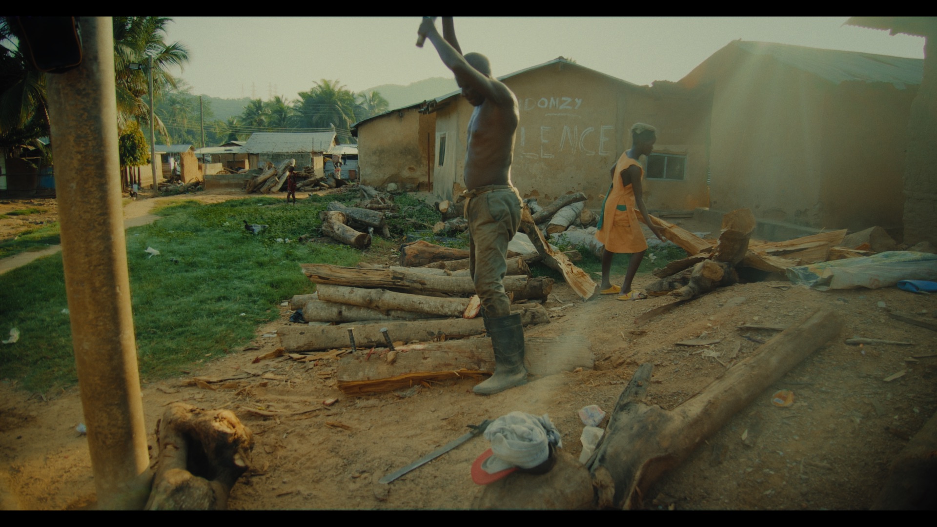 A man holding an item above his head stands on a dirt path beside a pile of logs in a rural area. A child in an orange dress walks in the background towards a building with a thatched roof.
