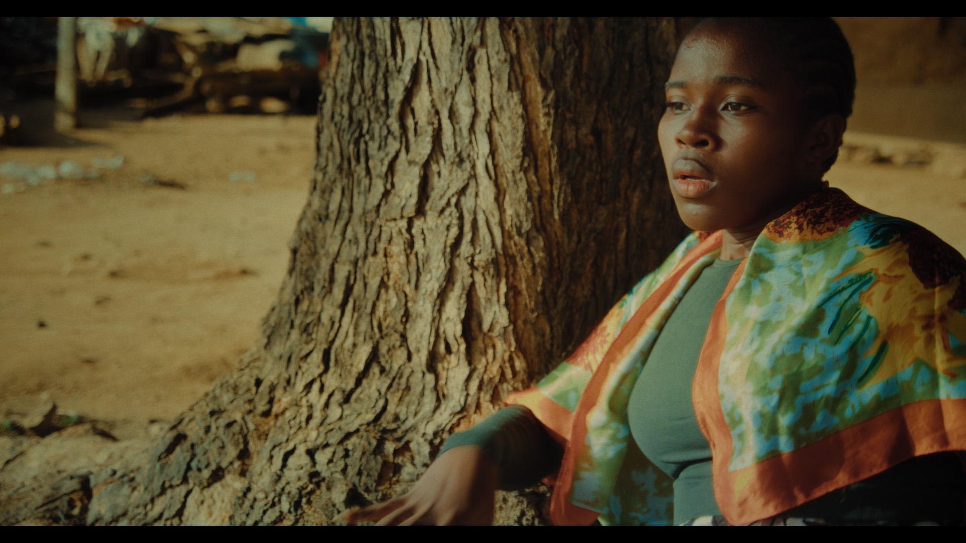 A young woman sits against a large tree trunk, wearing a grey top and a multicoloured patterned shawl. The background features an earthy, blurred landscape with natural light illuminating her face.