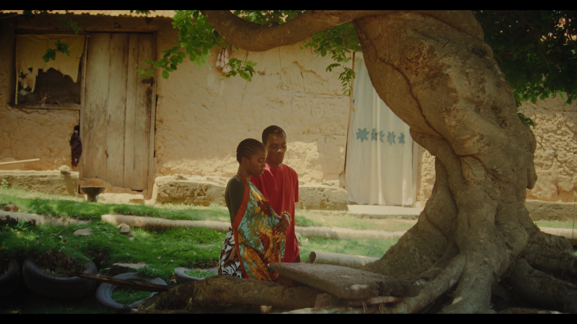 Two individuals stand near the base of a large tree in front of a traditional building with a weathered door. One person wears a colourful patterned dress, while the other is dressed in a red garment. The background features natural greenery.