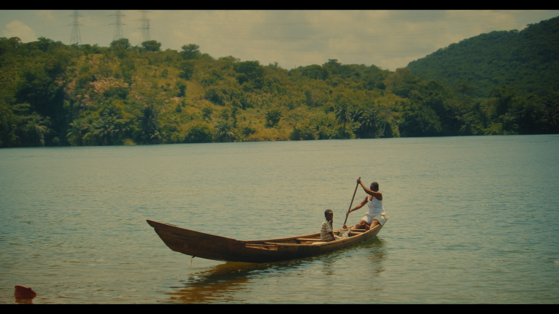 A wooden canoe floats on a calm river, with two individuals inside. One person navigates with a paddle while the other sits, surrounded by lush green hills and trees in the background.