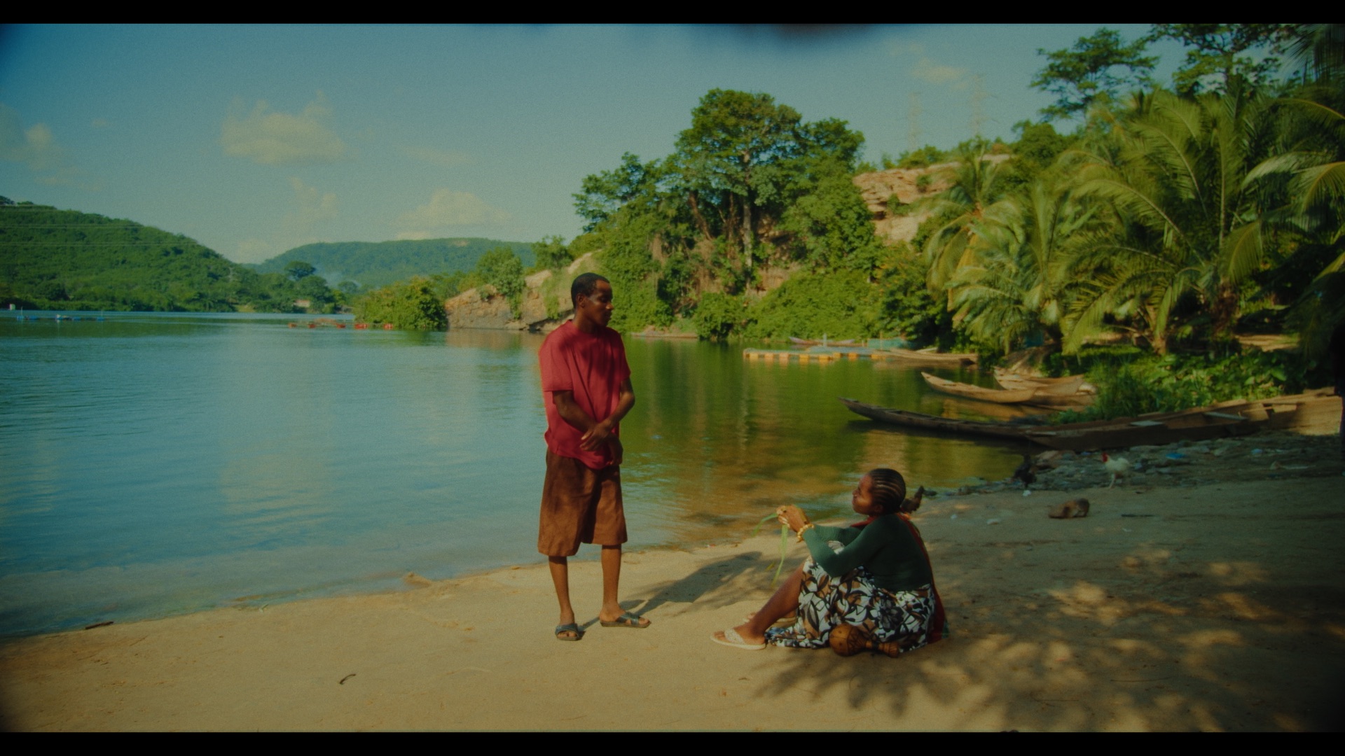 A man in a red shirt and beige shorts stands on the sandy shore next to a woman in a green top and patterned skirt, who is sitting on the ground. The setting includes a calm water body and lush green hills in the background.