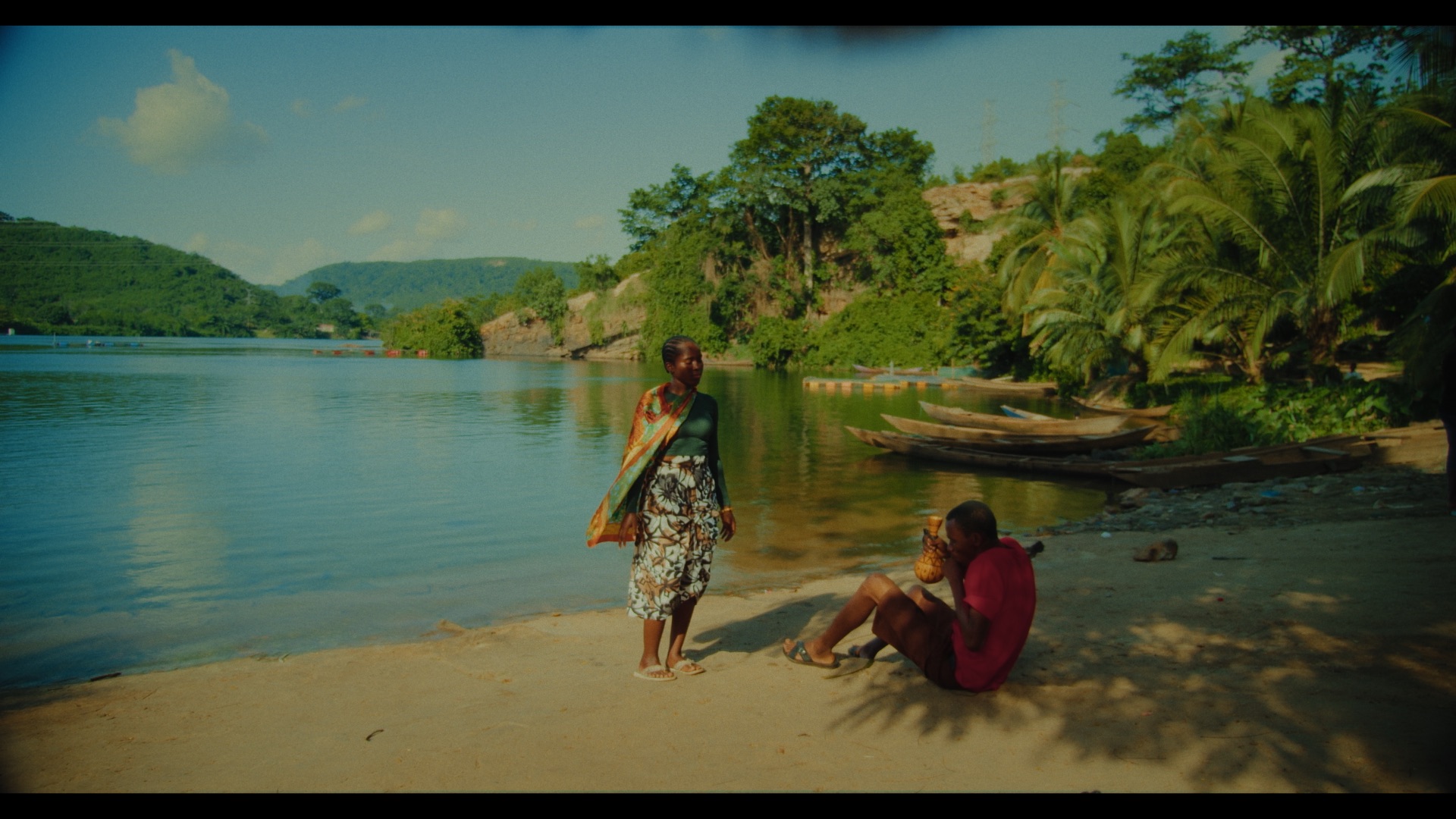 A woman in a patterned dress stands on a sandy beach near a lake, facing a seated man in a red shirt. The background features lush green hills, palm trees, and wooden canoes along the shore, under a clear blue sky.