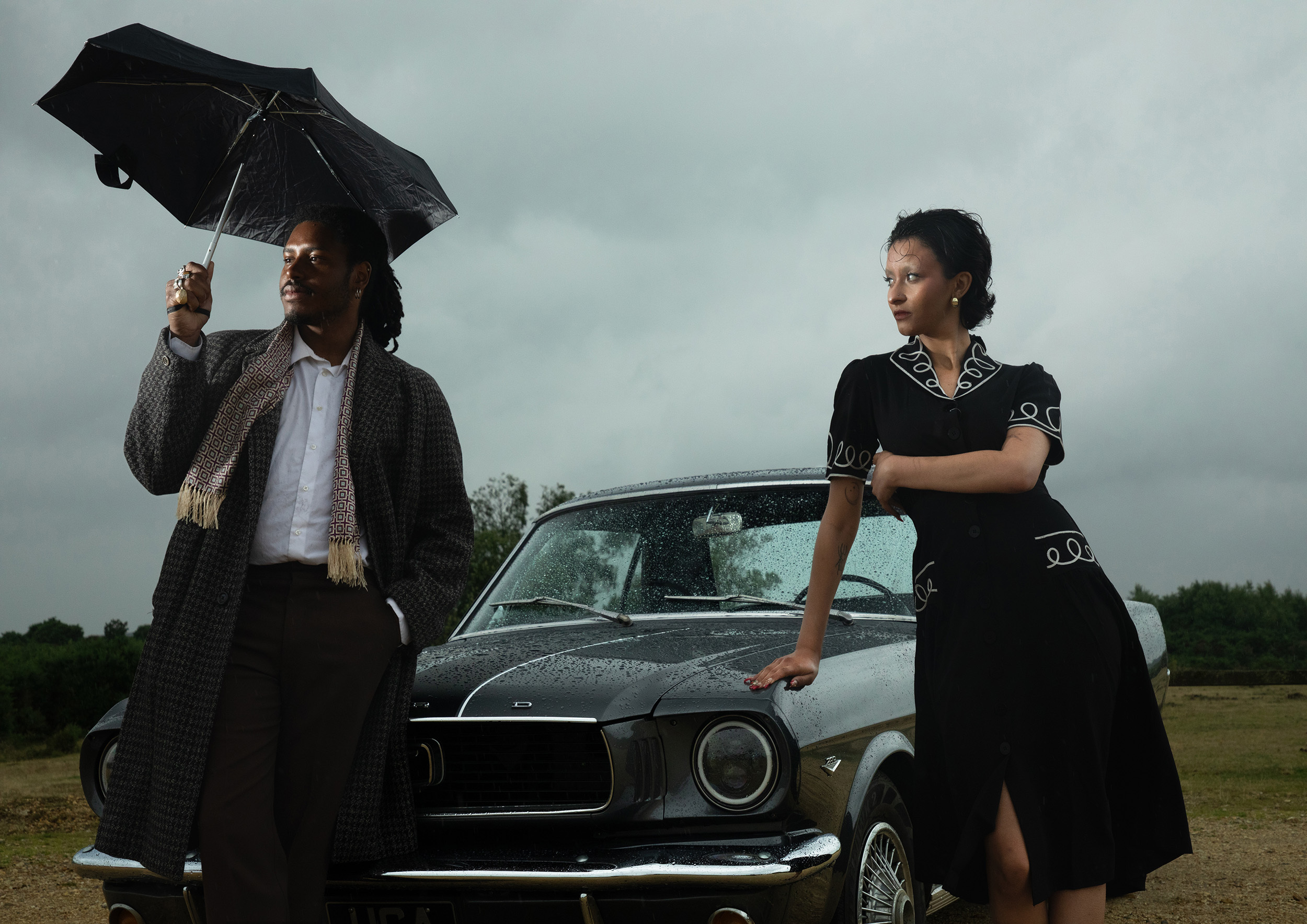 Two models leaning on a vintage car in the rain, the male-presenting model looks away under the protection of an umbrella, while the female-presenting model looks over to him with her arm wrapped around her chest