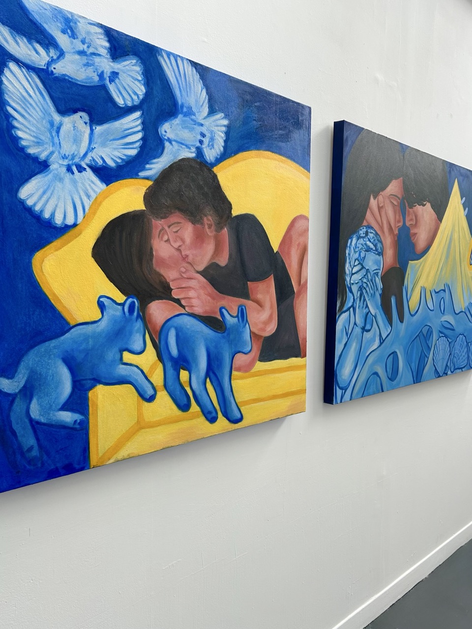 Two large paintings displayed on a white wall. The left painting features a couple on a yellow couch, surrounded by blue cats and white birds. The right painting displays two figures touching foreheads, with abstract blue shapes and yellow elements in the background.