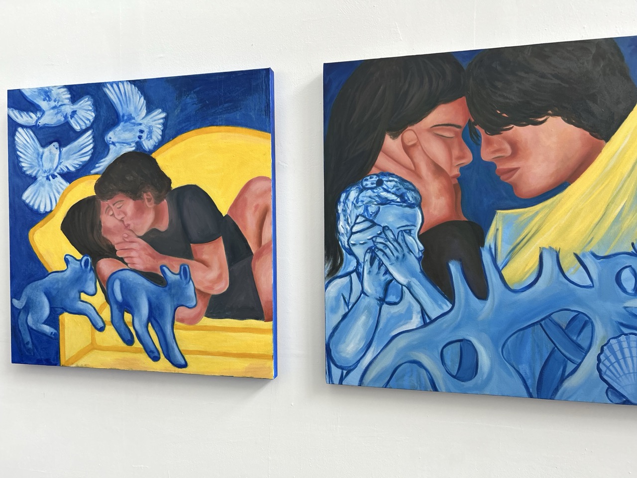 Two paintings displayed on a white wall. The left painting features a young man and woman resting together on a yellow couch, accompanied by blue cats and white doves in the background. The right painting depicts a close-up of the same couple's faces, surrounded by blue coral-like shapes.