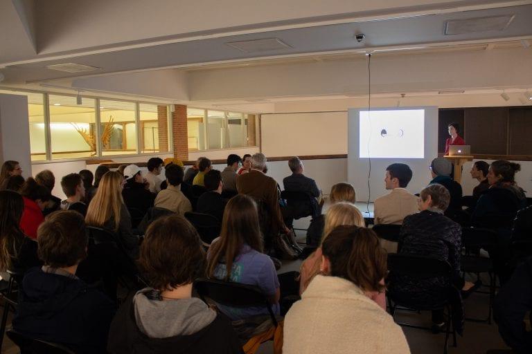 A crowded and dimly lit lecture theatre; a person at the front delivers the lecture with a bright screen to their side.
