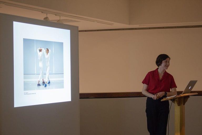 Alexandra Davenport stands at a lectern with a laptop open in front of them and a screen behind them, giving a lecture.