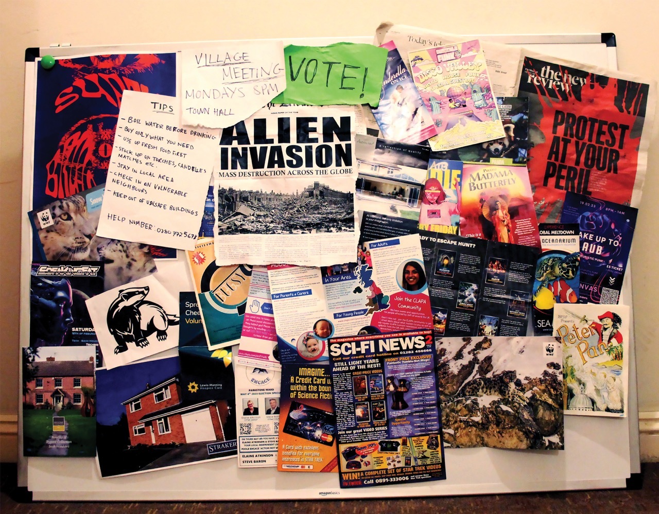 A large town hall notice board filled with leaflets and posters, with a newspaper front cover cut out in the centre, reading “Alien Invasion”.
