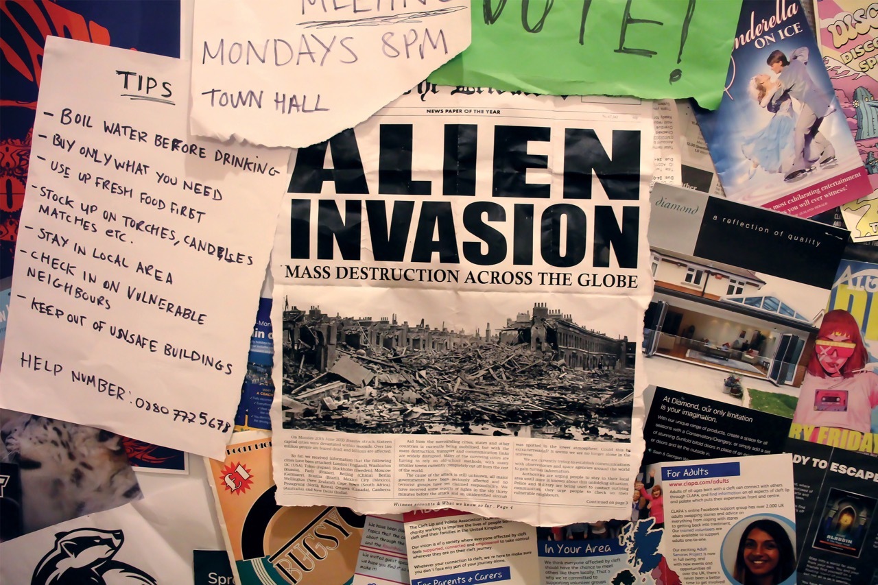 Close-up of town hall notice board, focusing on the newspaper cut out, reading “Alien Invasion – Mass destruction across the globe”.