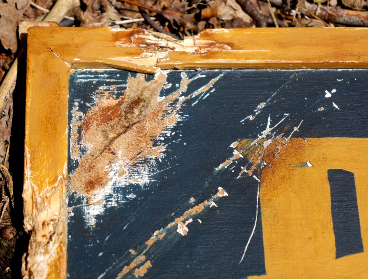 Close-up of the top left-hand corner of the sign, it is scratched and splintered, as if a bullet has grazed it.