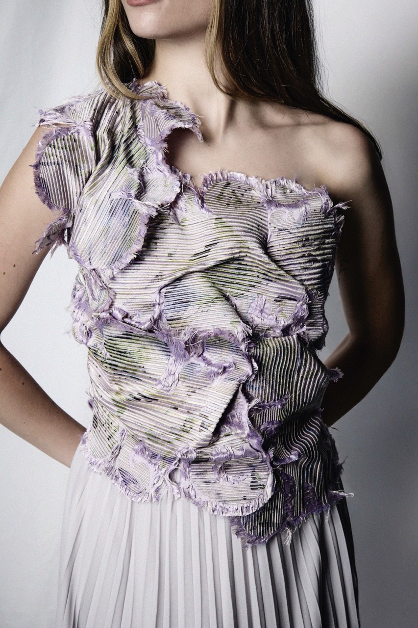 Woman wearing one-shoulder dress with purple ruffled textured bodice and white pleated skirt against grey background.