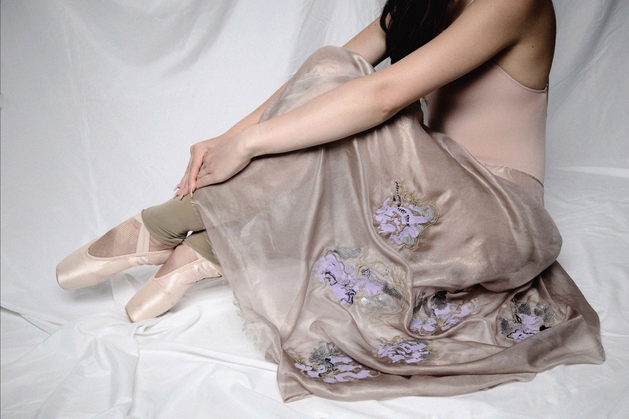 Woman in beige dress with purple floral embroidery sitting on white fabric, wearing ballet pointe shoes with ribbon ties.