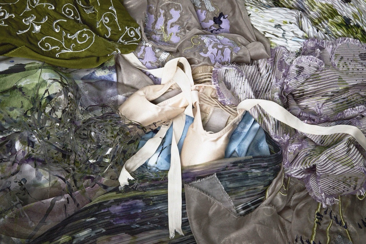 Pale pink ballet shoes with cream ribbons resting on crumpled fabric in green, purple, and blue tones with floral patterns.