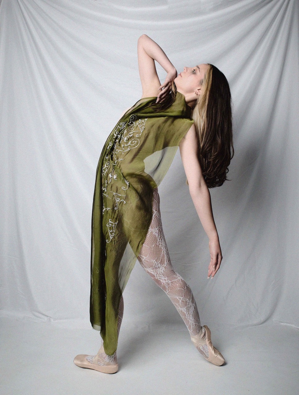 Woman in olive green embroidered dress and nude tights poses with head tilted back, one arm raised, against white backdrop.