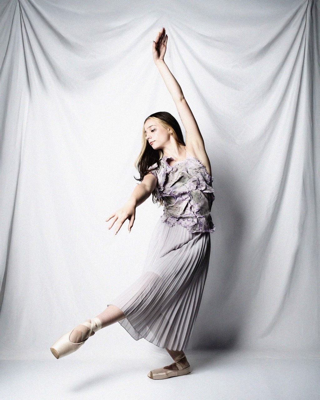 Woman in grey off-shoulder top and pleated skirt dancing with one arm raised, against white fabric backdrop.