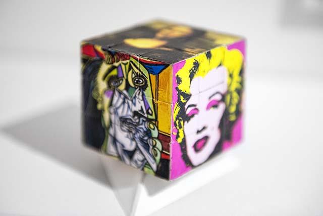 A cube sculpture showing three pieces of artwork. Da Vinci's Mona Lisa appears on the top face. The Weeping Woman by Picasso is on the left-hand face, and part of The Marilyn Diptych by Andy Warhol is on the right-hand face.