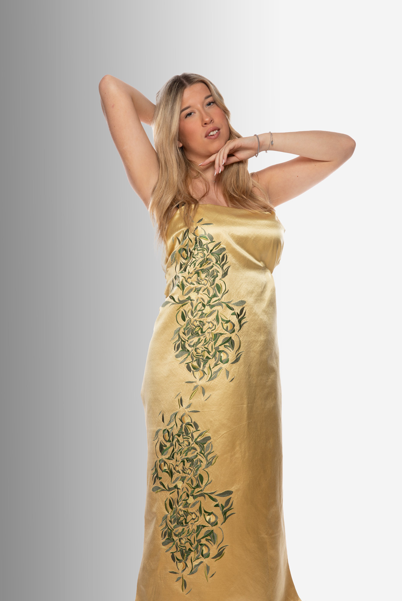 Blonde woman in golden satin dress with green floral embroidery, hands positioned behind head, against white background.
