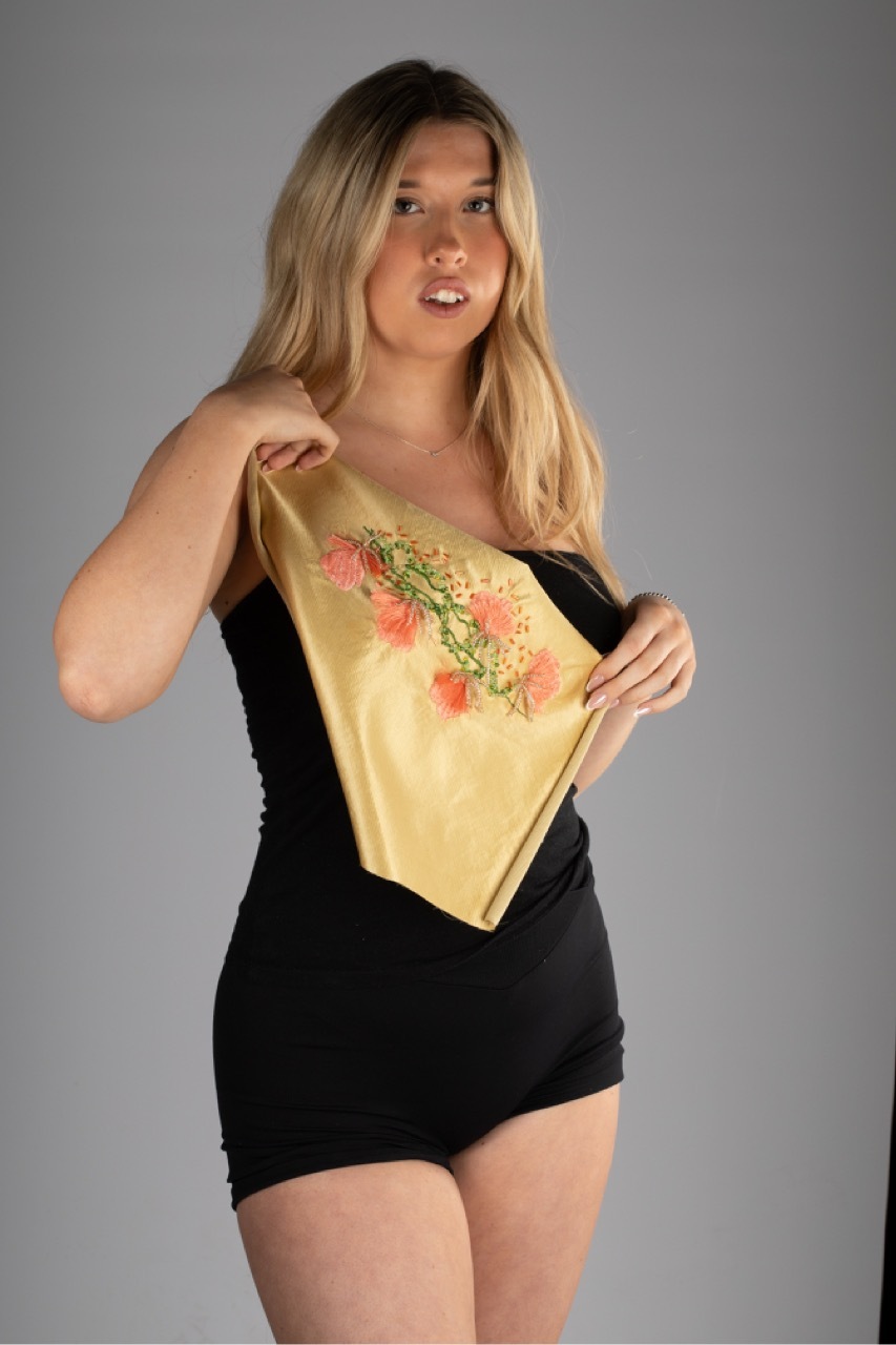 Woman with blonde hair holding yellow silk scarf with pink floral embroidery, wearing black dress against grey background.