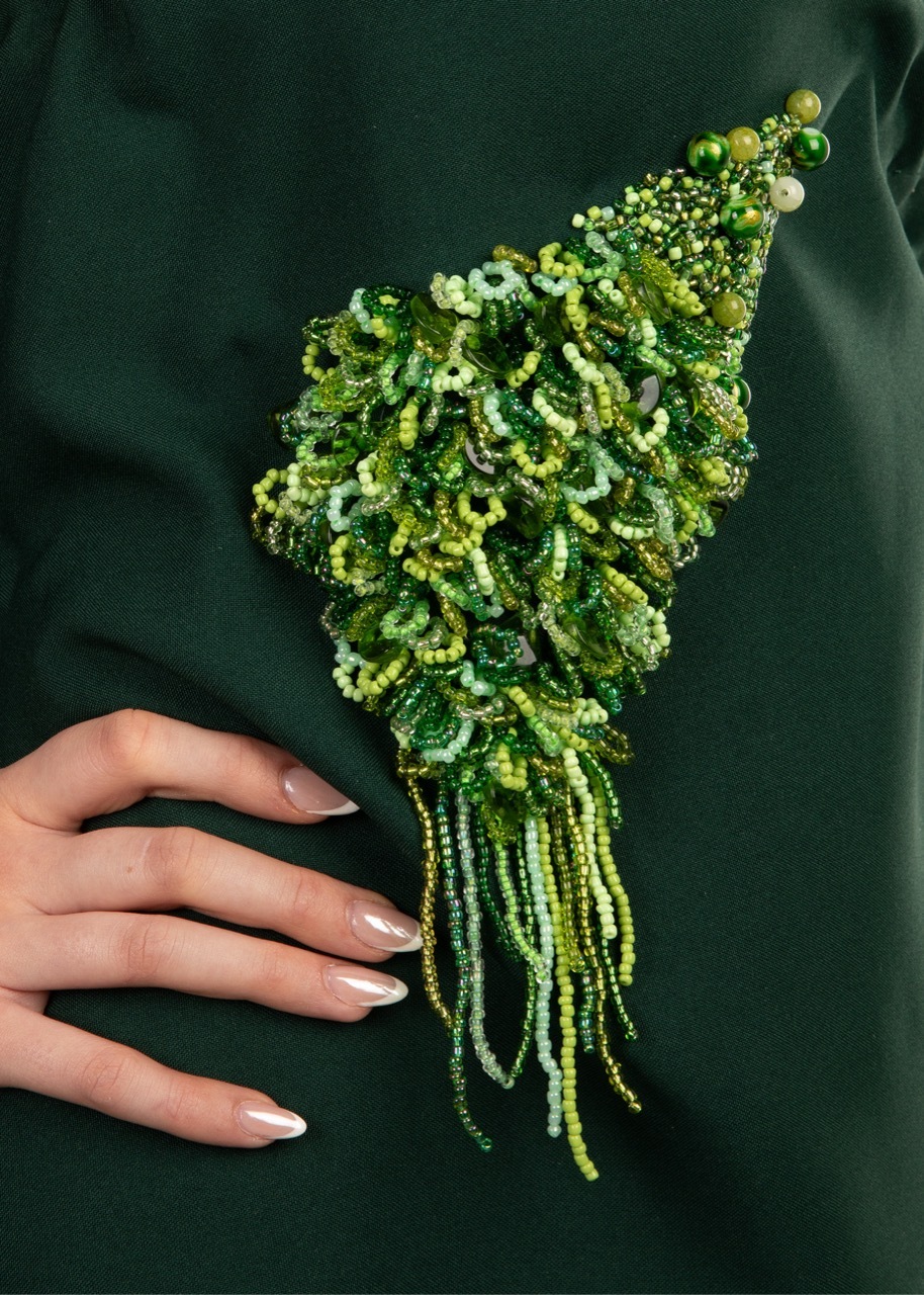 Hand with white nail polish beside elaborate green sequined and beaded decorative piece with dangling fringe on dark green fabric.