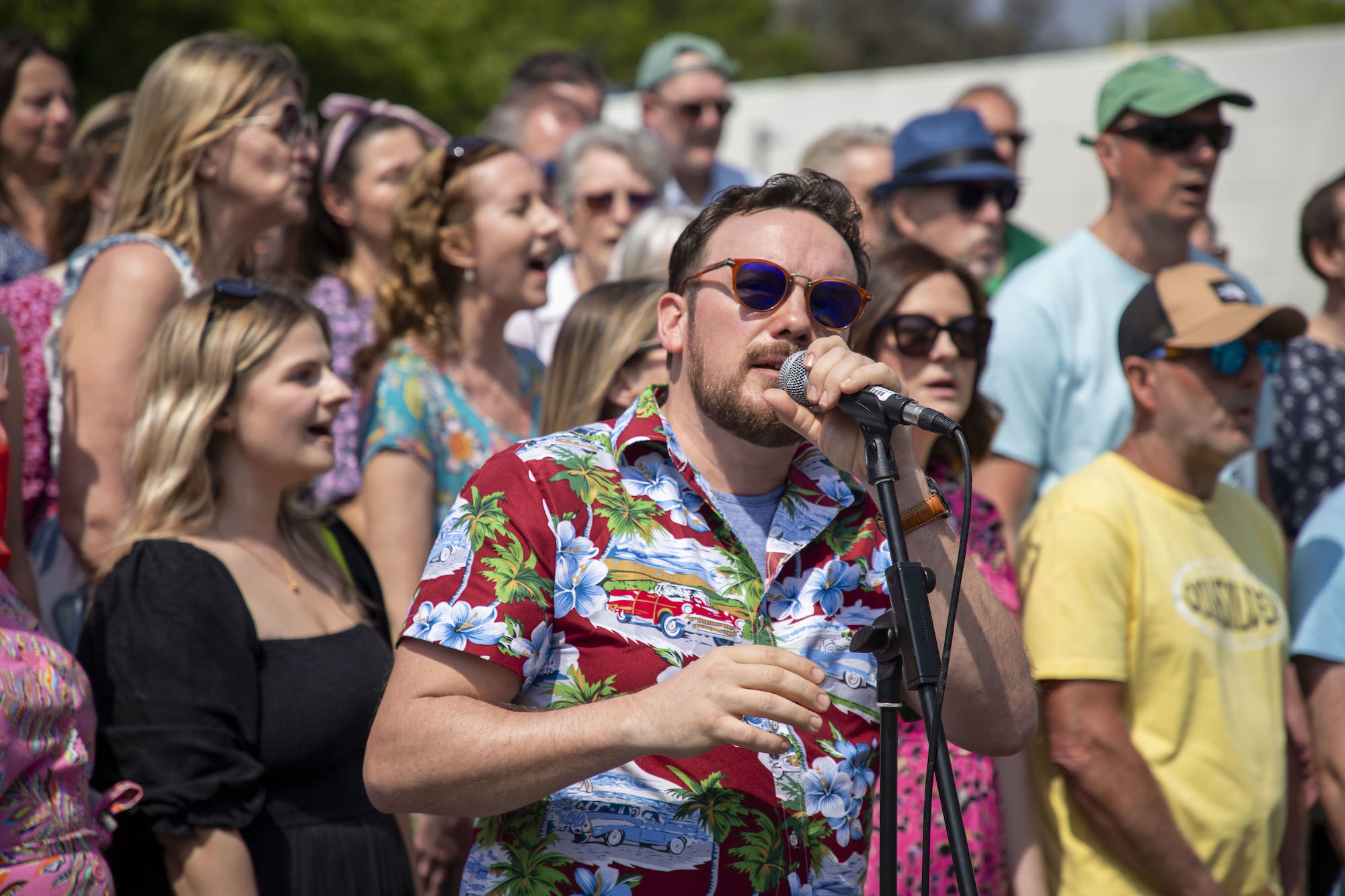 A performer speaking or singing into a microphone, with a large crowd behind him, at the 2024 Alice in Spring event in Bournemouth.