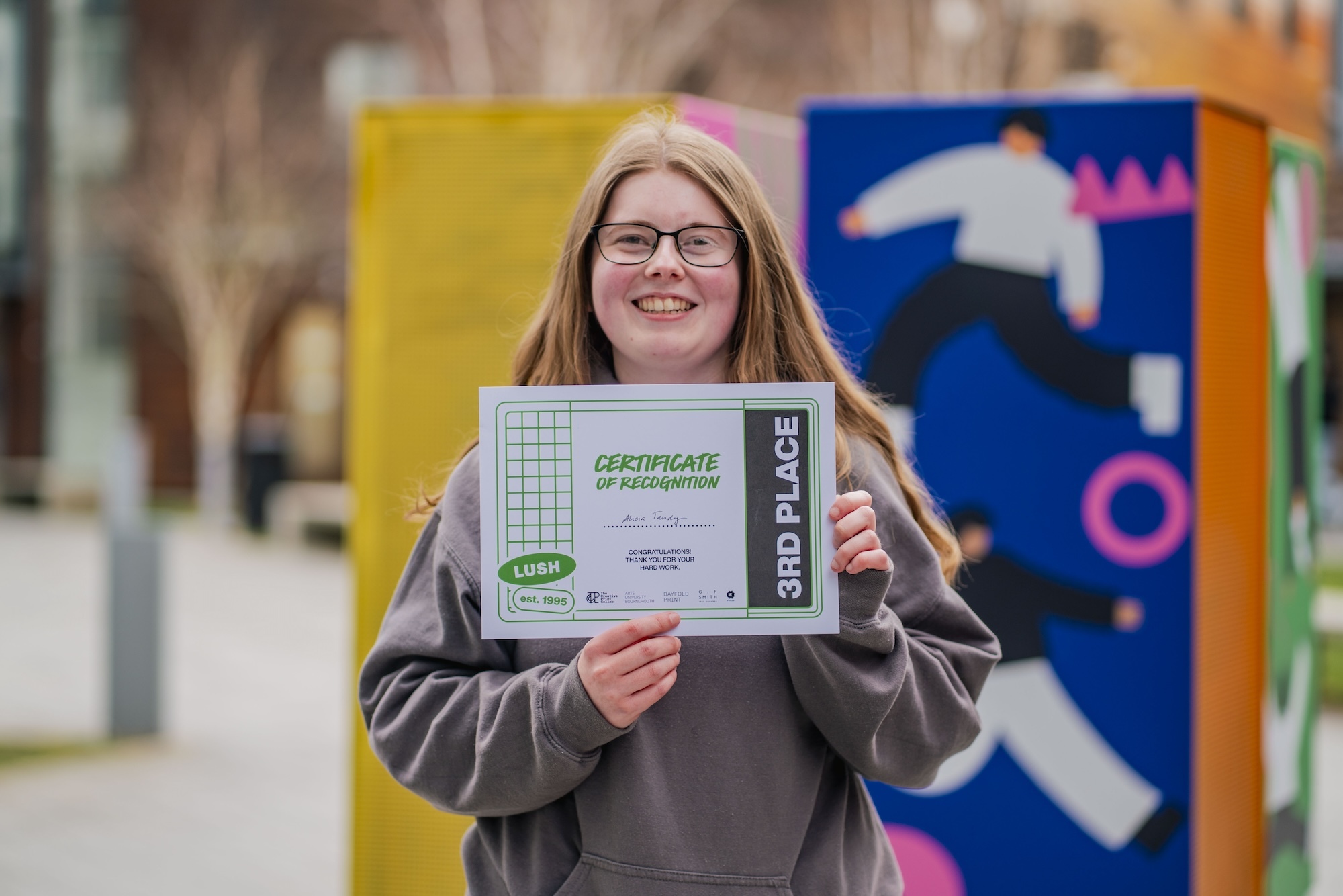 BA (Hons) Graphic Design student A. Tandy, holding a certificate for third place, posing for a photo in the courtyard outside University House, Arts University Bournemouth.