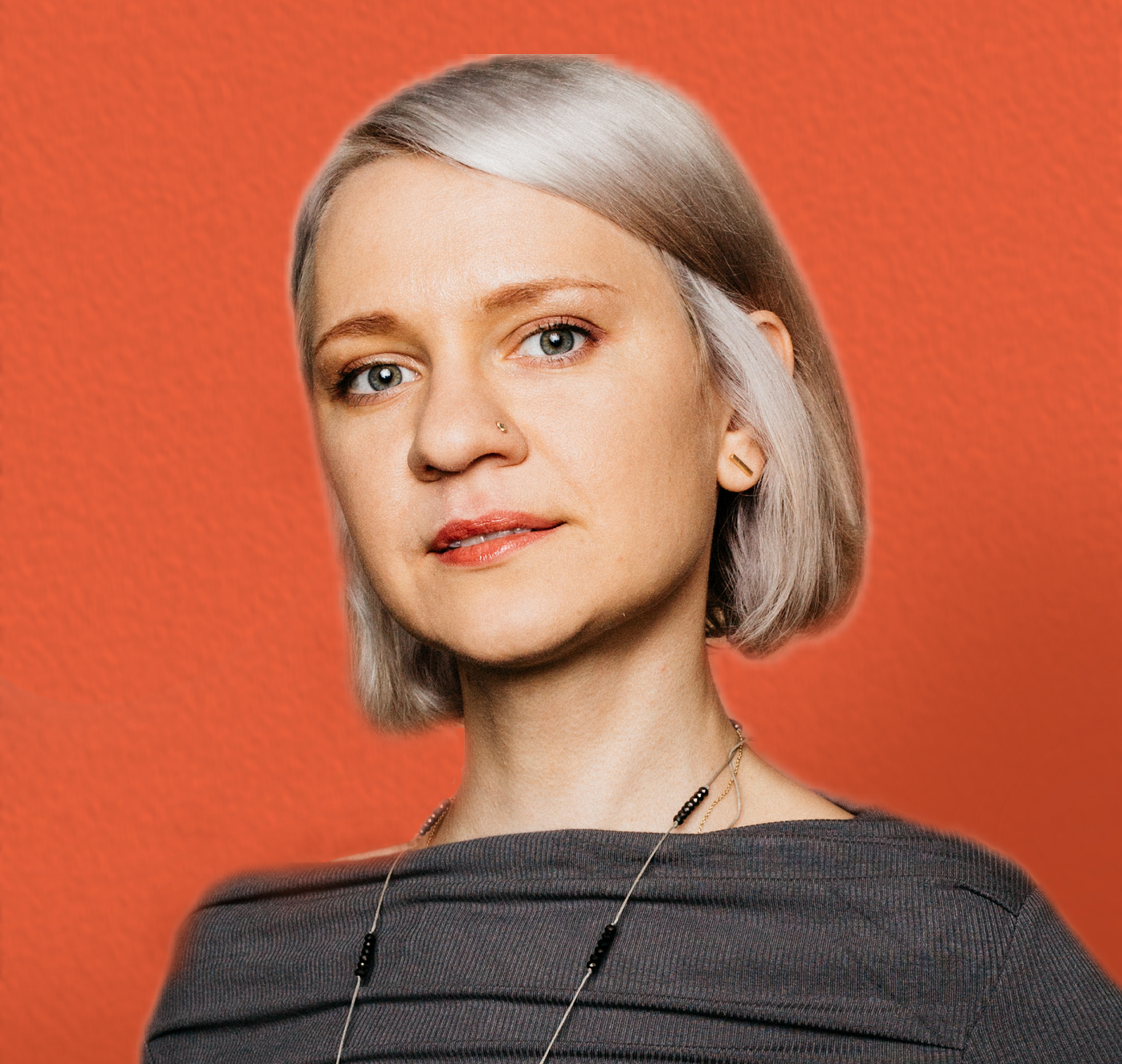 Woman with blonde bob haircut wearing grey off-shoulder top and necklace against bright orange background.