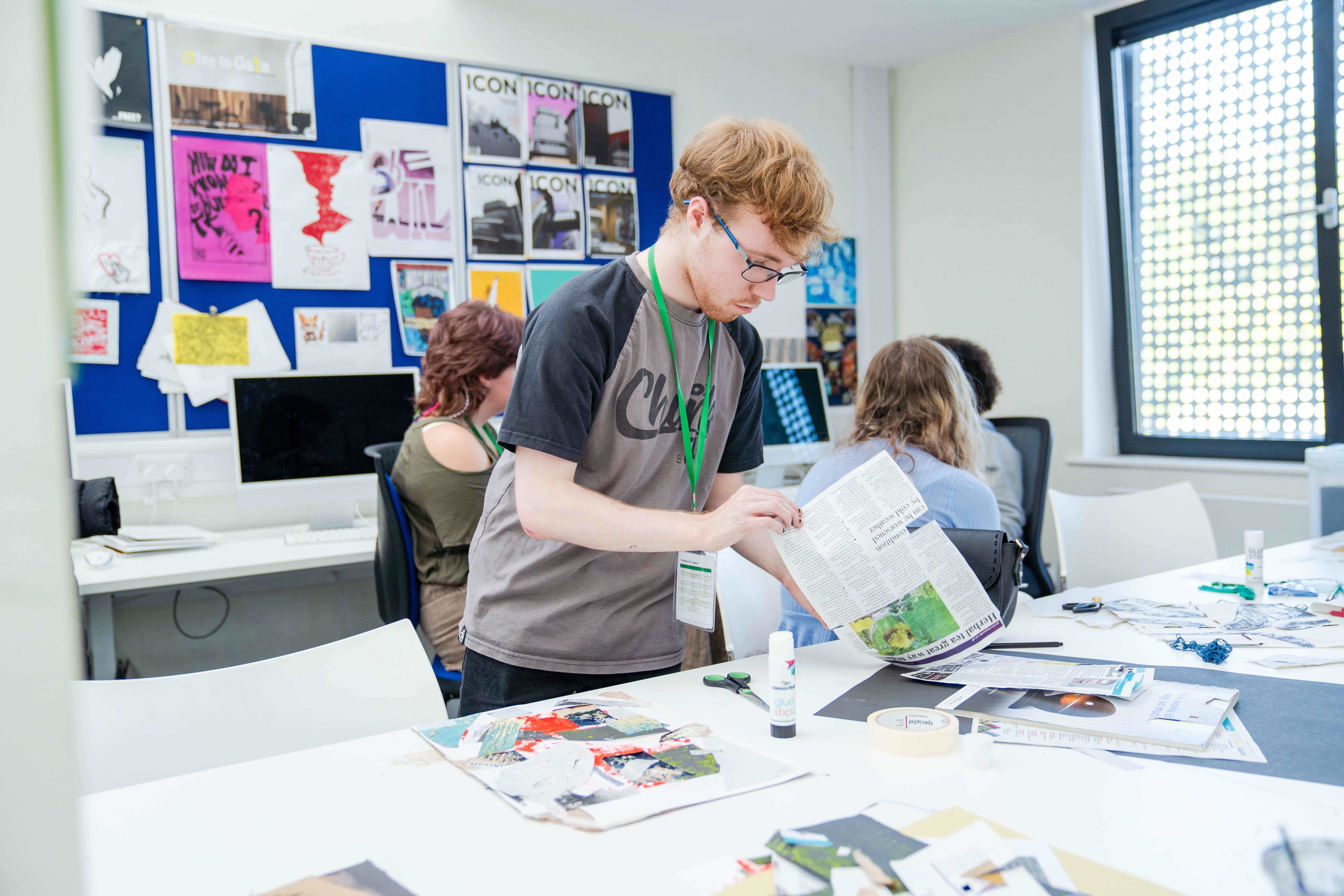 A student is working at a desk holding a page of a magazine in the middle of making a collage.