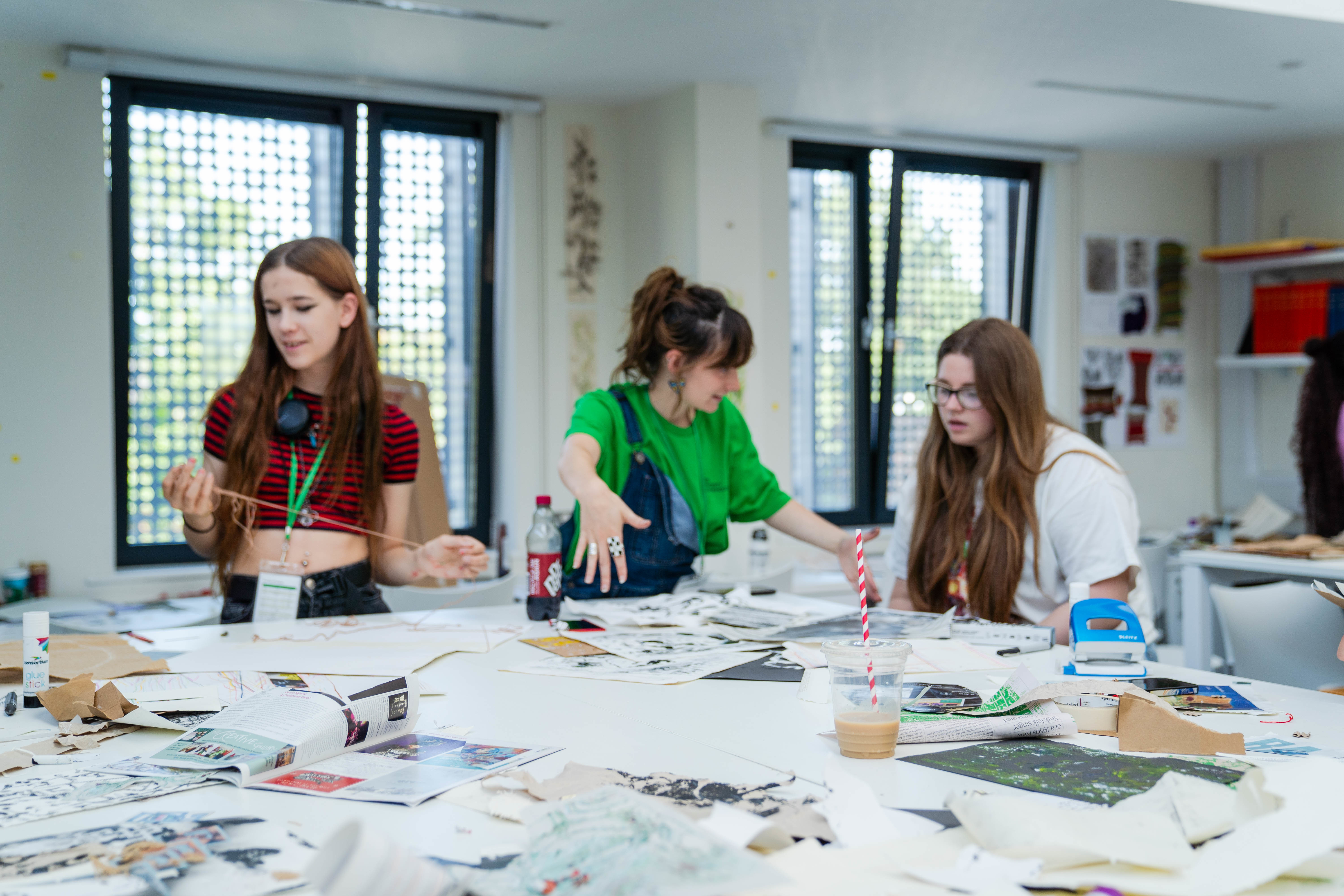 An AUB Outreach Ambassador standing between two young people in a seminar room, around a large table covered with magazine clippings.
